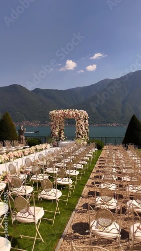 Aerial drone view of luxury destination wedding at Lake Como, Italy. White aisle, floral arch, and neat chairs set against breathtaking lake and mountain scenery