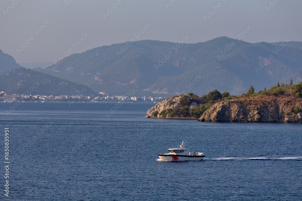 Obraz premium A boat is sailing on blue water near a rocky coast. Mountains are visible in the distance with a hazy sky overhead in Icmeler - Marmaris - Mugla - Turkey