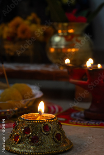 A beautifully lit decorative diya sits in focus during a traditional Indian pooja, surrounded by offerings, lamps, and flowers.