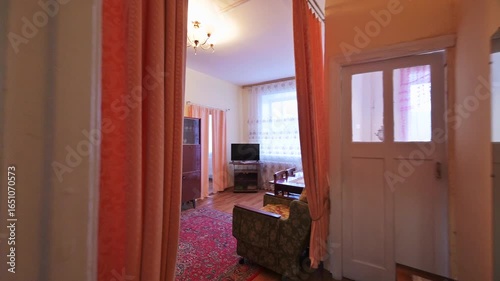 vintage-style living room with orange curtains, a patterned rug, and classic furniture. An open doorway reveals a glimpse into the warm interior