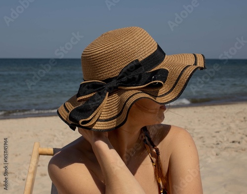 A red-haired woman in a hat with a bow, from under which only her lips are visible, sits on the beach