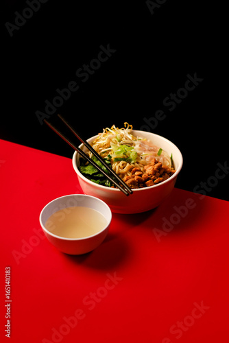Chicken noodle bowl with vegetables, bean sprouts, and scallions served with clear soup and chopsticks on red and black background. Asian food.