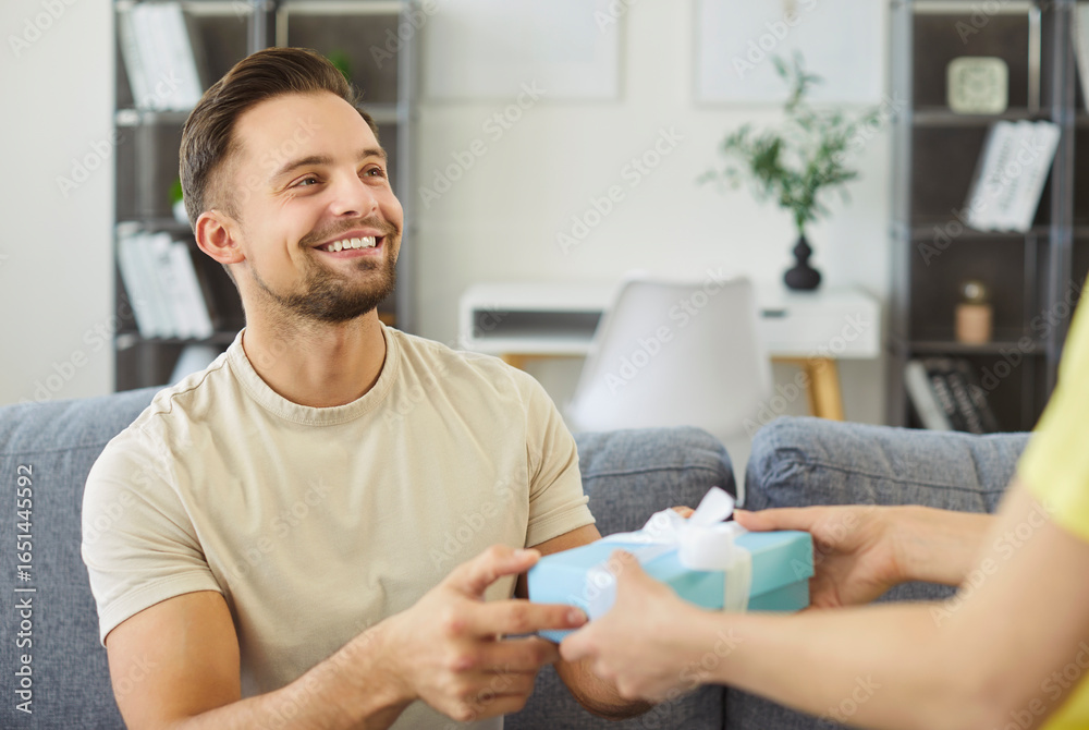 Fototapeta premium Young woman giving present gift box congratulating her smiling boyfriend sitting on sofa in living room at home. Happy couple celebrating holiday together. Love and Valentines day concept.