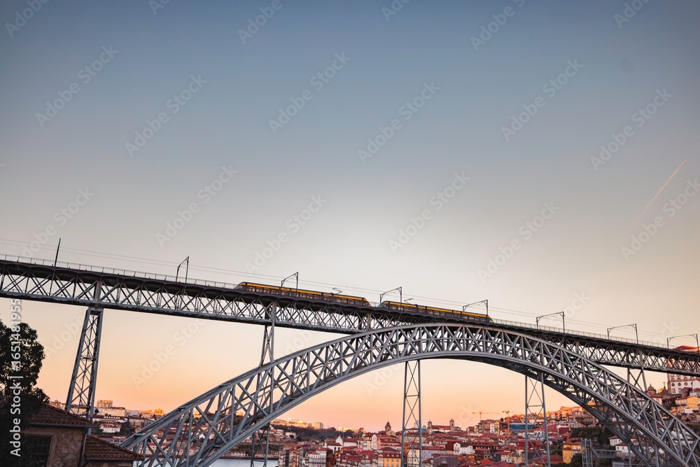 Fototapeta premium A yellow train crosses the Dom Luis I Bridge, gracefully spanning the Douro River. The sun descends, casting a warm, vibrant glow over the cityscape of Porto, Portugal