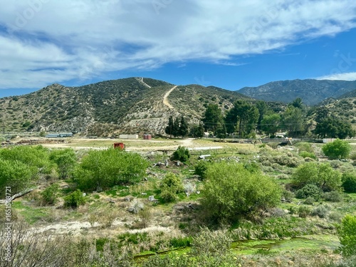 Rolling hills with dirt roads, green vegetation in foreground, red buildings, mountains in background under cloudy blue sky in arid landscape near Acton, California