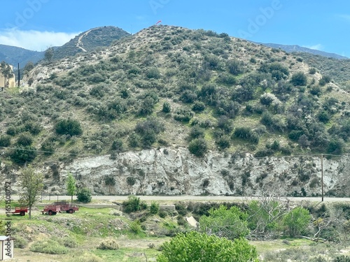 Green hillside with exposed rocky cliff face, red farm vehicles parked below, power lines visible, lush vegetation under clear blue sky near Acton, California