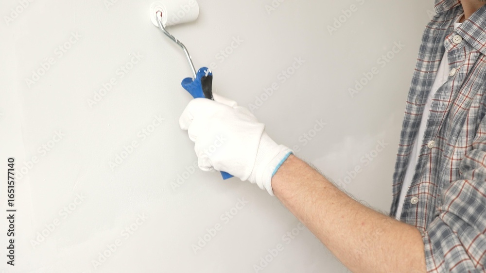 Naklejka premium Male painter's gloved hand gripping paint roller, spreading white paint across wall during home interior transformation with professional precision, close-up view. Renovation concept