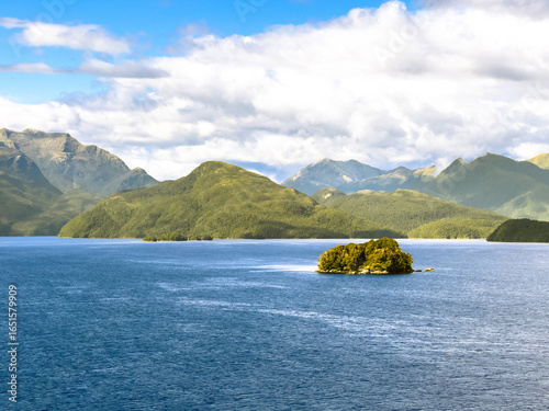 Serene Milford Sound with a small lush island in crystal-clear blue waters, framed by green mountains and a bright cloudy sky, New Zealand