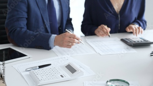Photos Calculator and magnifying glass over financial documents opposite financial team analyzing data, reviewing reports, collaborating at modern office workspace, close up view