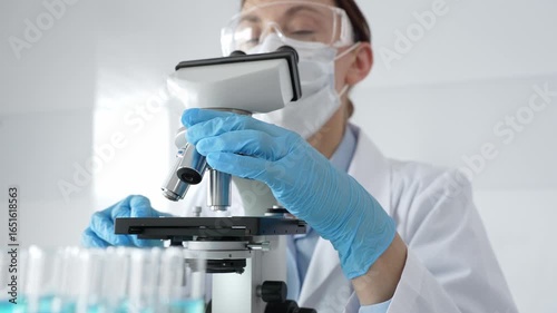 Female scientist wearing protective equipment checking microscope, analyzing blue test tubes in modern laboratory research setting. Medicine, healthcare and science concept