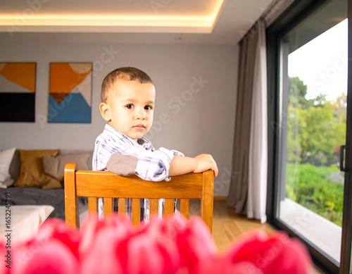 A toddler sits in a wooden chair, looking back at the camera.  Blurred pink flowers are in the foreground