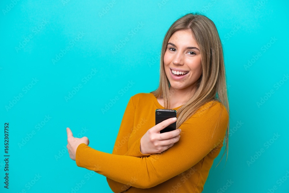 Young caucasian woman isolated on blue background using mobile phone and pointing back Young caucasian woman isolated on blue background using mobile phone and pointing back
