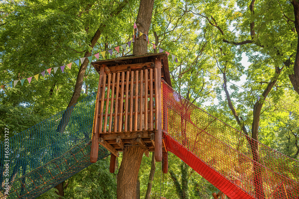 Naklejka premium A wooden treehouse with colorful rope bridges and safety nets surrounded by lush green trees in a sunny outdoor playground.