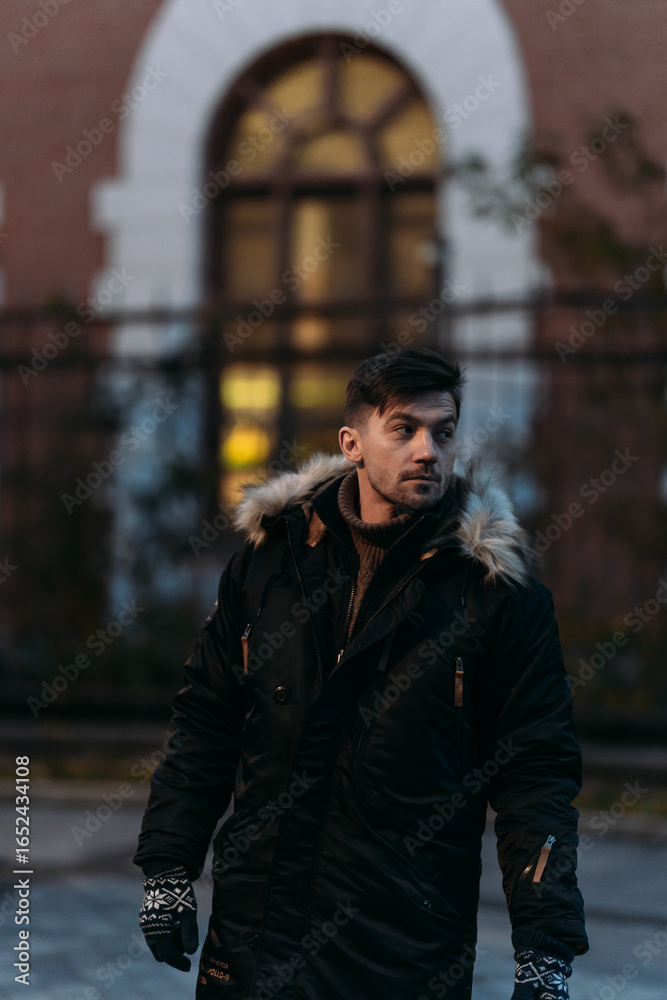 Fototapeta premium Man in black winter coat with fur hood and gloves standing outdoors near brick building. Warm clothing, urban autumn fashion, casual walk, modern city style, architectural background