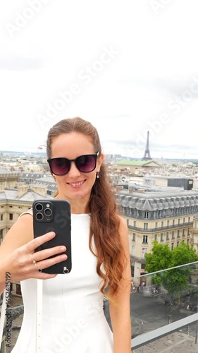 Stylish young woman streaming video on smartphone from rooftop in Paris with Eiffel Tower