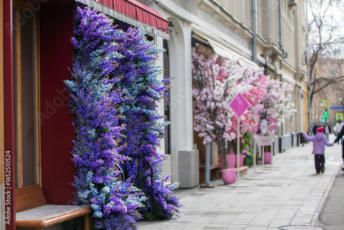 Colorful floral decorations adorn a city street, with vibrant purple and pink blossoms lining storefronts, creating a charming urban spring scene