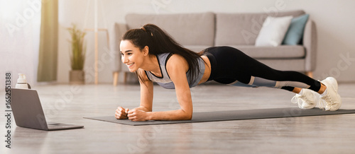 Sporty millennial woman planking in front of laptop, living room interior, side view, copy space. Healthy young lady in sportswear exercising at home, watching online fitness class