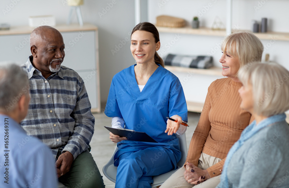 Obraz premium Friendly pretty young woman in blue workwear psychologist having conversation with multiracial group of senior people sitting in circle, psychological support for senior people at nursing home