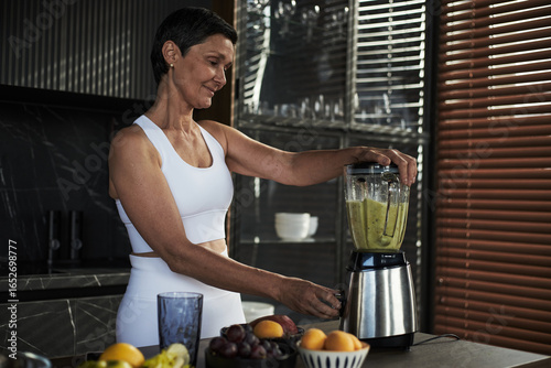 Fotografi Middle aged Caucasian woman blending smoothie in kitchen, smiling while preparin
