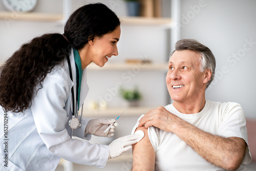 Millennial doctor making medical drug injection or anti-covid vaccine to disabled senior man in wheelchair at home. Nurse immunizing senior impaired male patient, doing shot of antibiotic indoors