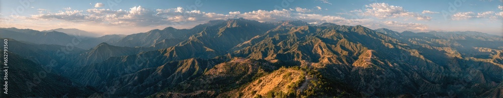 Naklejka premium Panoramic view of a mountain range at sunrise or sunset. Misty, layered peaks stretch across the image. Soft light bathes the landscape