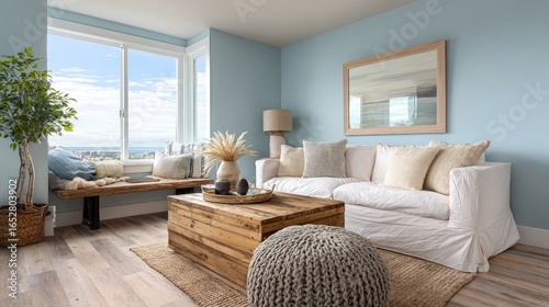 Cozy living room with large windows, a sofa made of wood and white cushions, a knitted rug on the floor, a sea view outside the window, a picture frame hanging above the couch, neutral blue walls, coa