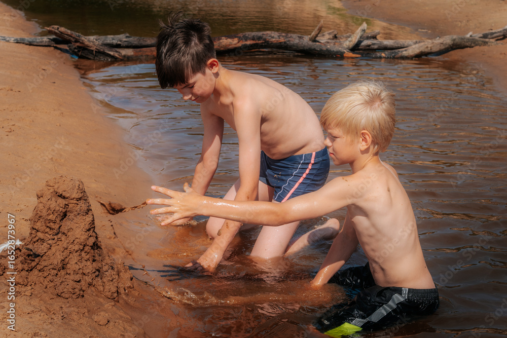 Fototapeta premium Shirtless boys play in muddy water, building a long mud structure on a sandy riverbank in bright summer sunshine. A playful and creative outdoor scene.