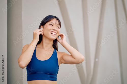 Fotografie Happy asian sportswoman wearing a blue sports bra and adjusting her wireless ear