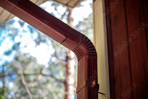 Detail view of a red downspout corner attached to a wooden cabin, with trees and sky blurred in the background.
