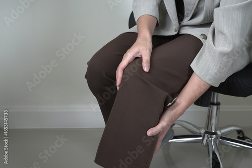 Asian businesswoman suffers from painful severe knee joint pain sitting on a chair in office and massaging leg muscles, knee injury, overworked, illness, office syndrome, cramps or rheumatism concept.