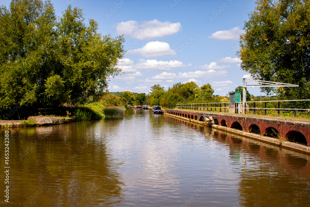 Fototapeta premium Grafton Regis Northamptonshire England UK 08 16 2025 Weir crane and narrowboats on a tree lined Grand Union Canal above River Tove showing low water levels challenging navigation in summer drought