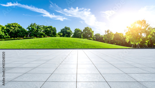 Fototapeta Naklejka Na Ścianę i Meble -  Serene park landscape: expansive paved plaza, lush green hill, vibrant trees under a bright summer sky, idyllic outdoor scene.