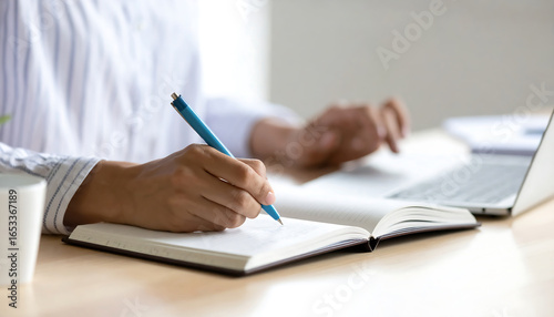 Close-up of a person's hand writing in a notebook near a laptop, showcasing a calm and focused work-from-home atmosphere, ideal for productivity, planning, and journaling concepts.