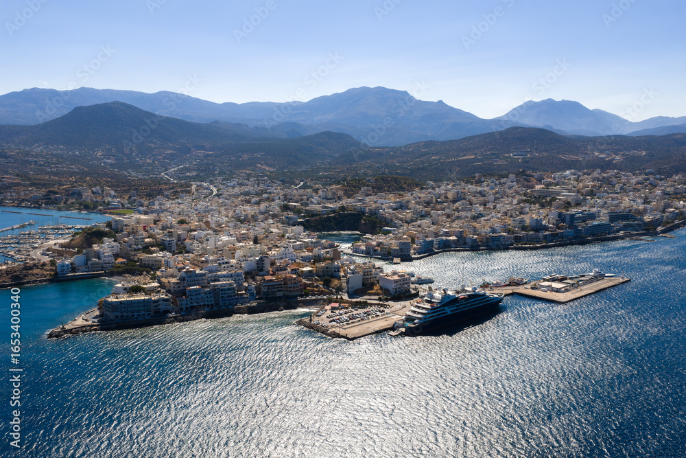 Obraz premium Aerial view of Agios Nikolaos, Crete, showing the harbor with a large yacht, smaller boats, white and pastel buildings, and surrounding mountains.
