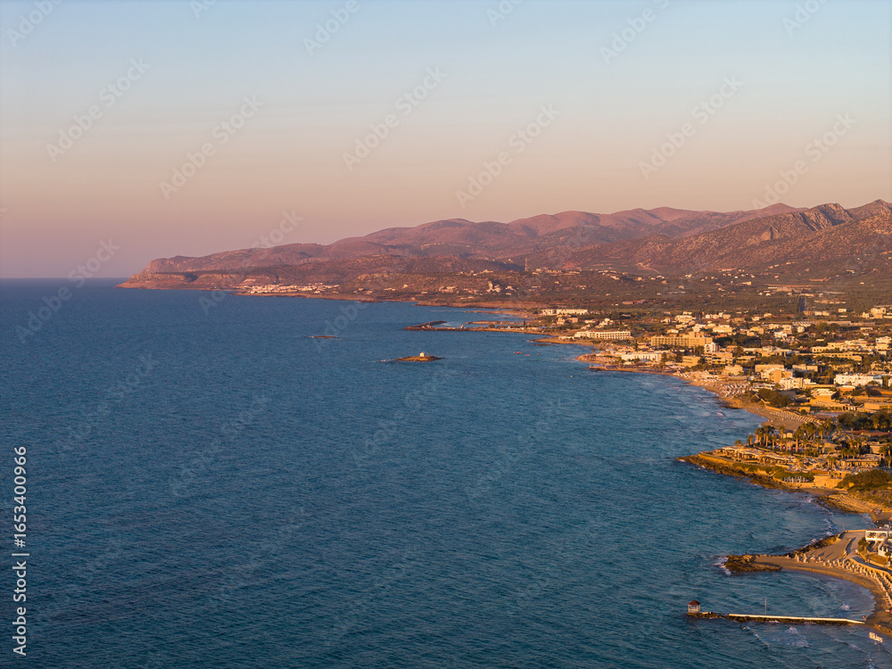 Fototapeta premium Aerial view of Stalida, Crete, showing the Aegean Sea, rugged coastline, small beaches, and town buildings with hills and mountains at sunset.