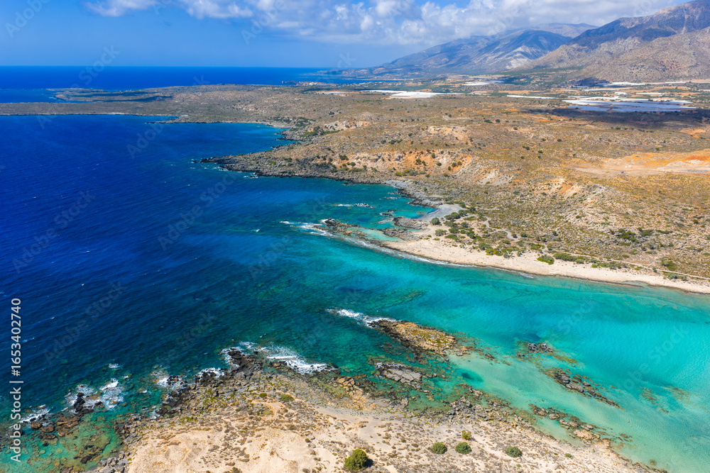 Obraz premium Aerial view of Elafonisi Beach in Crete, Greece, showing turquoise and blue waters, shallow lagoons, rocky outcrops, and distant mountains under clouds.