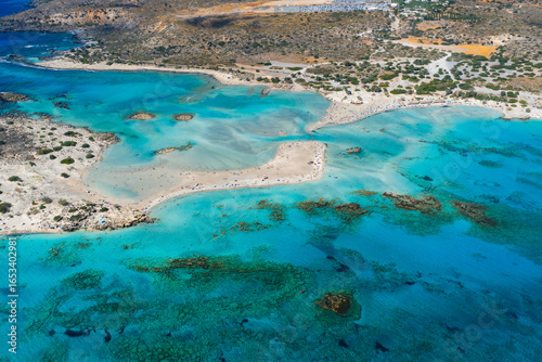 Fototapeta Naklejka Na Ścianę i Meble -  Aerial view of Elafonisi Beach in Crete, Greece, showing turquoise waters, pinkish sand, shallow sandbars, rocky islets, and sparse vegetation.