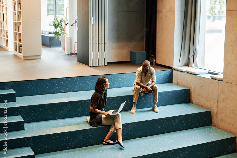 © TrueFrame Collective - Horizontal high angle view long shot of young African American man and Caucasian woman sitting on stair bench in modern library using Internet, drinking coffee and chatting © TrueFrame Collective - Horizontal high angle view long shot of young African American man and Caucasian woman sitting on stair bench in modern library using Internet, drinking coffee and chatting