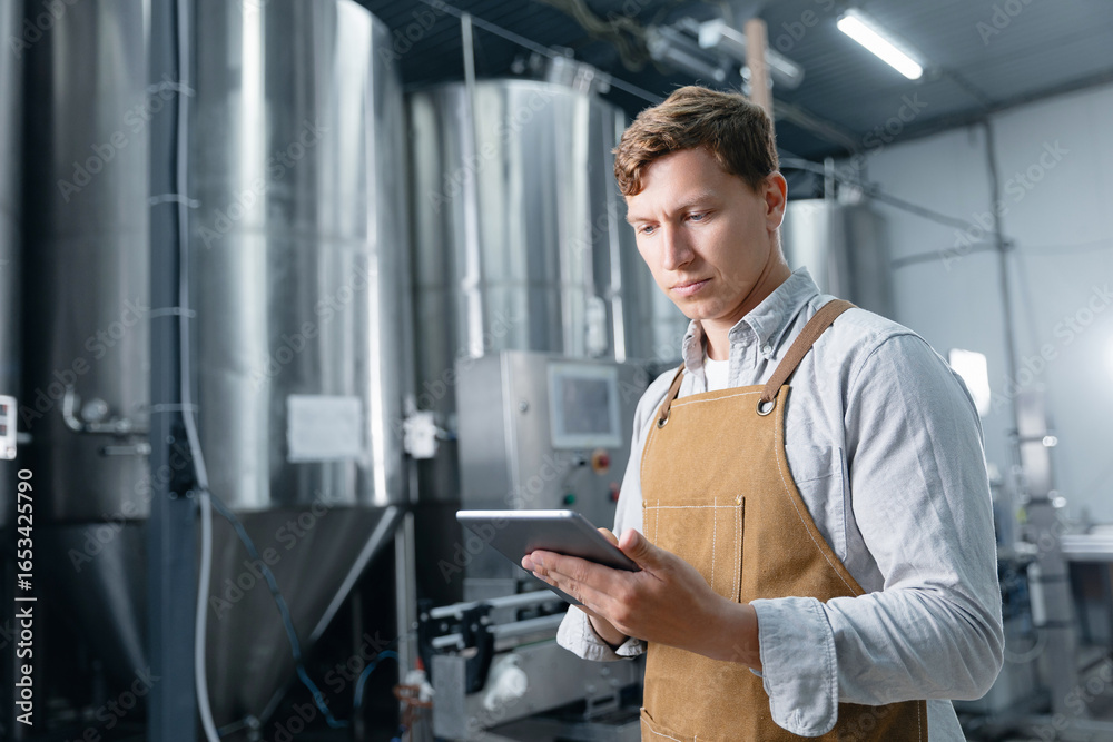 Fototapeta premium Male brewer using tablet in modern brewery with large steel tanks