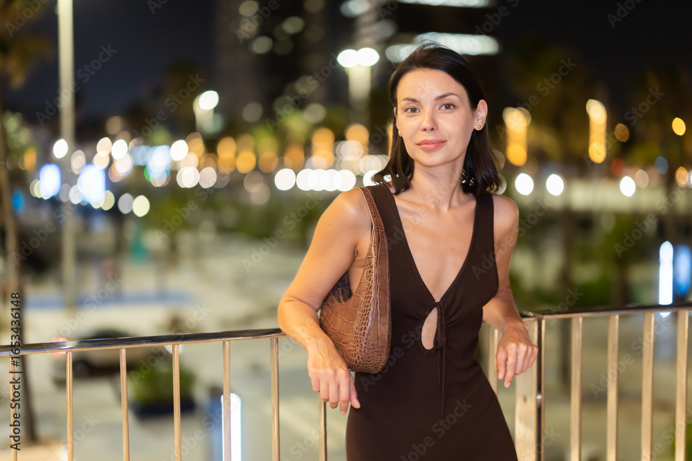 Fototapeta premium Happy young woman leaning on metal railing against background of blurred city lights of Barcelona