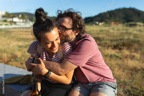 A man embraces his partner with love, while they spend time at the sun, sitting on a walk, in a coastal area, while drinking beer