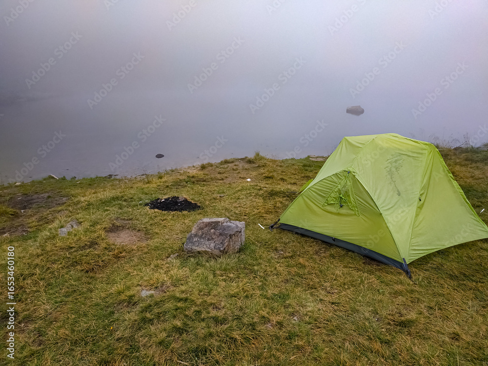 Obraz premium Green tent on misty lake shore with a fireplace nearby. Foggy camping scene, mysterious atmosphere, outdoor adventure in nature.