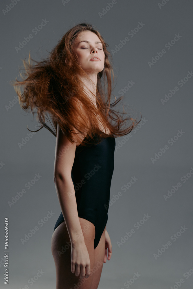 Fototapeta premium Young woman in black bodysuit with flowing hair posing confidently against gray background.