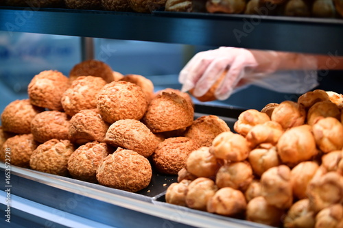 Professional bakery worker wearing gloves while handling fresh cream puffs. Popular French-origin pastry with crispy shell and creamy filling displayed in commercial bakery setting.