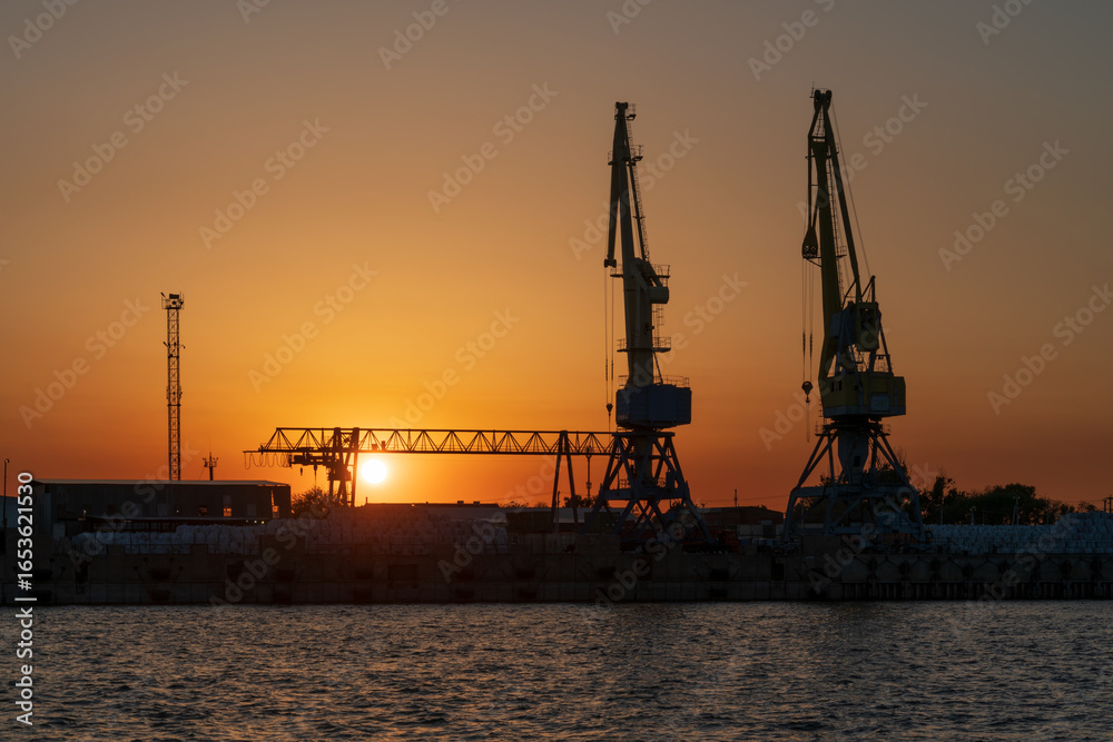 Obraz premium Port cranes for cargo transshipment on the mooring line of the Astrakhan port on the right bank of the Volga River against the background of the setting sun, Astrakhan, Russia