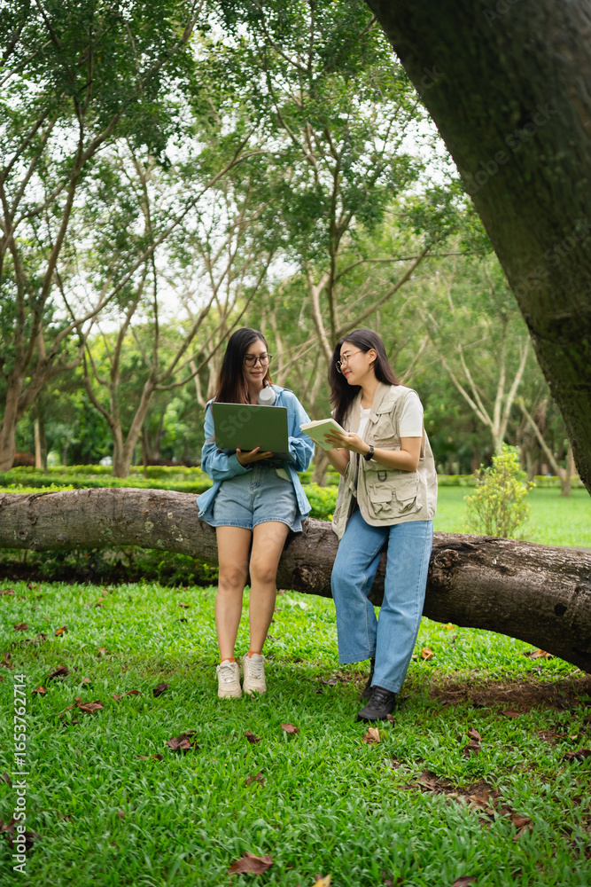 Fototapeta premium Two Young Women Collaborating Outdoors While Using Laptop and Reading Notes in a Lush Green Park Surrounded by Trees and Nature