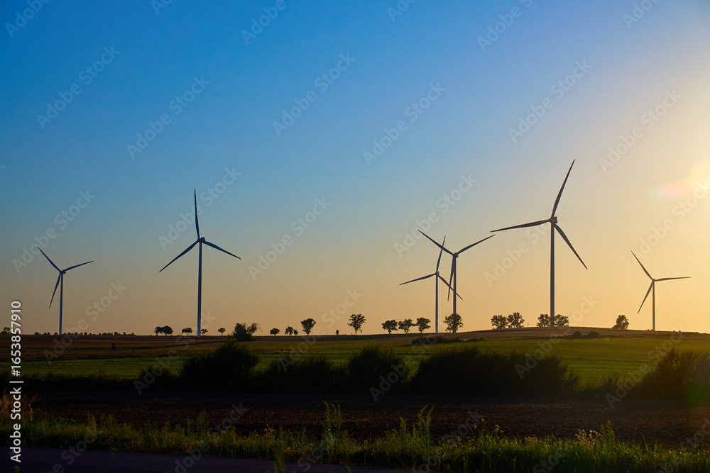 Wind turbines in open farmland with green fields and trees under evening sky. Windmills for production clean energy at sunset. Concept of renewable energy, sustainable resources and green technology Wind turbines in open farmland with green fields and trees under evening sky. Windmills for production clean energy at sunset. Concept of renewable energy, sustainable resources and green technology