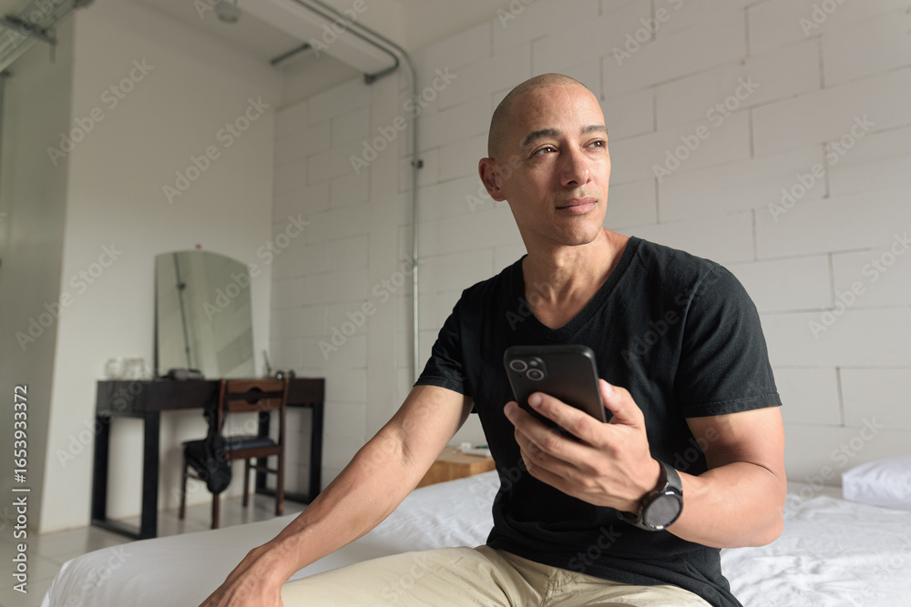 Fototapeta premium Bald Hispanic man sitting on bed at home using phone looking through window