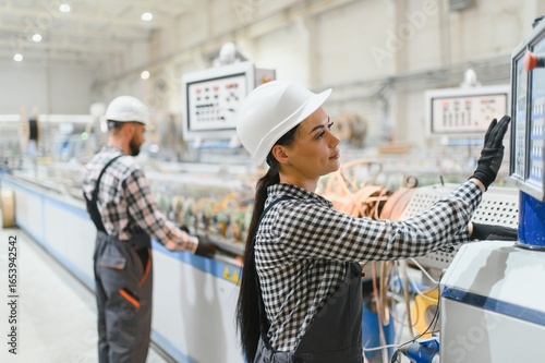 Slika na platnu Female factory worker operating machinery with male colleague in background