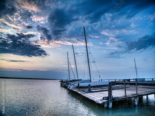 Peaceful sunrise at Breitenbrunn, Lake Neusiedl, Austria. Sailboats moored at a wooden pier with golden morning light reflecting on calm rippling water, symbolizing nature, sailing, and tranquility.